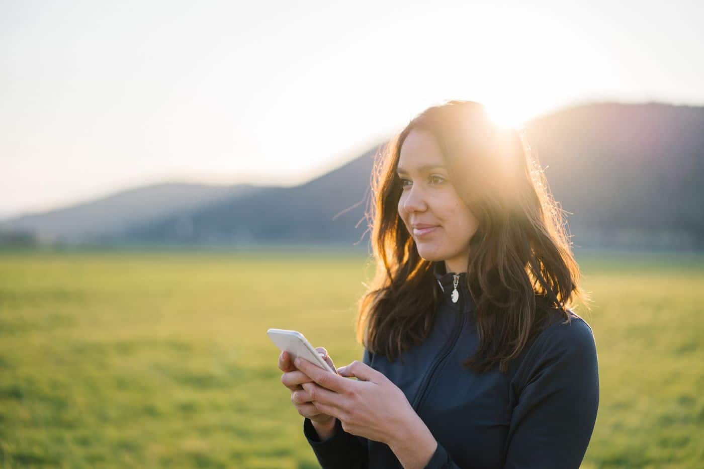 Woman outdoors smiling while looking at her phone in a sunlit field