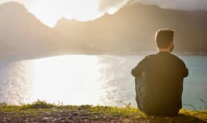 man sitting alone on the ground looking at a scenic view of a lake and mountains