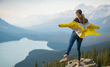 a scenic view of a mountain range, pine trees, and a body of water in the distance. a woman is balancing on a rock overlooking the view